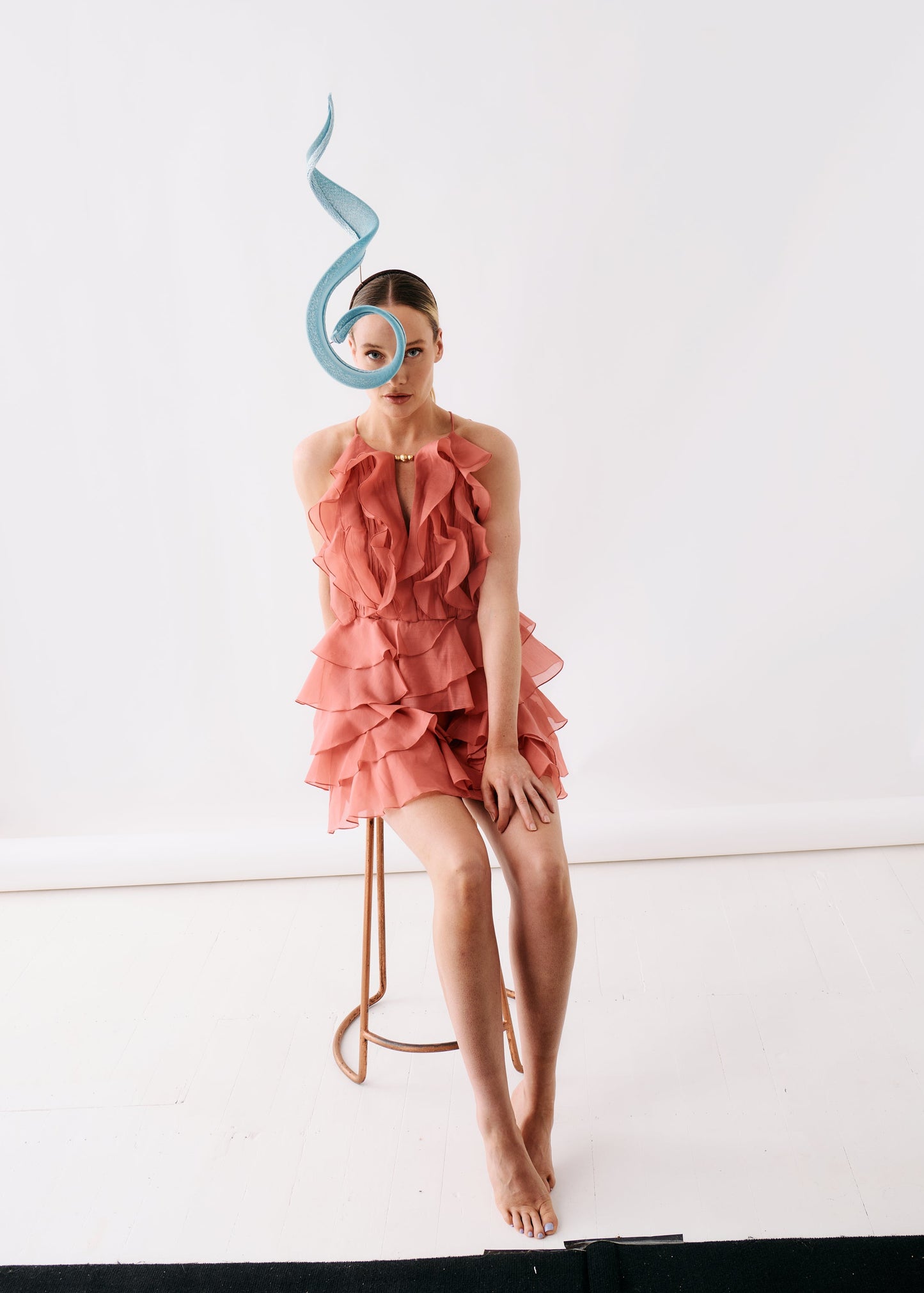 A model sits on a minimalist copper-toned stool in a studio setting, wearing a ruffled coral pink halter dress and a light blue sculptural headpiece.