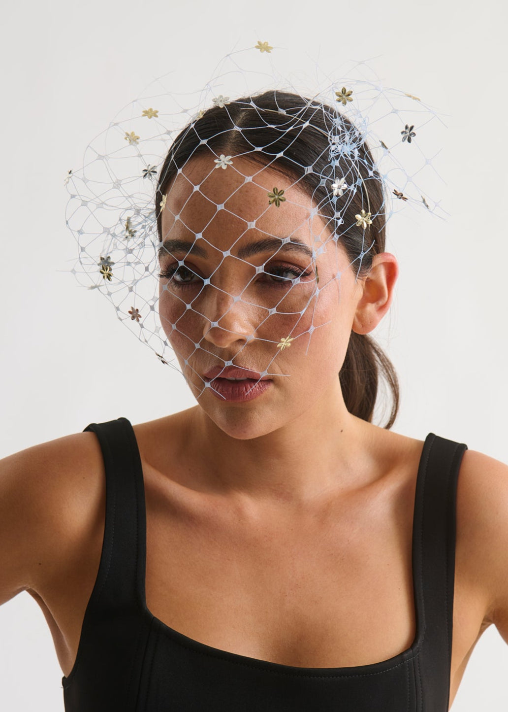 Woman wearing a black dress and a decorative veil headpiece with flowers against a plain background