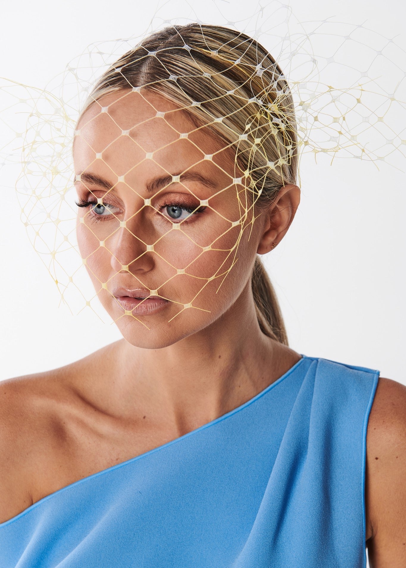 Woman wearing a blue one-shoulder dress with a netting headpiece on a white background