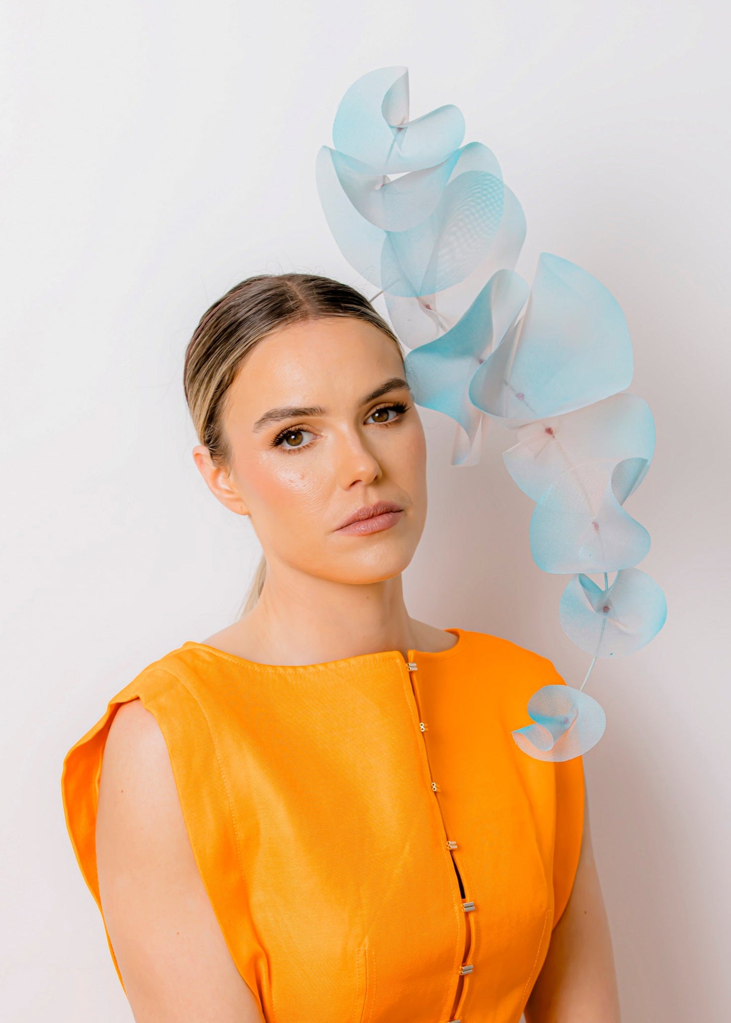 Woman in an orange dress with a blue ruffle headpiece against a white wall backdrop