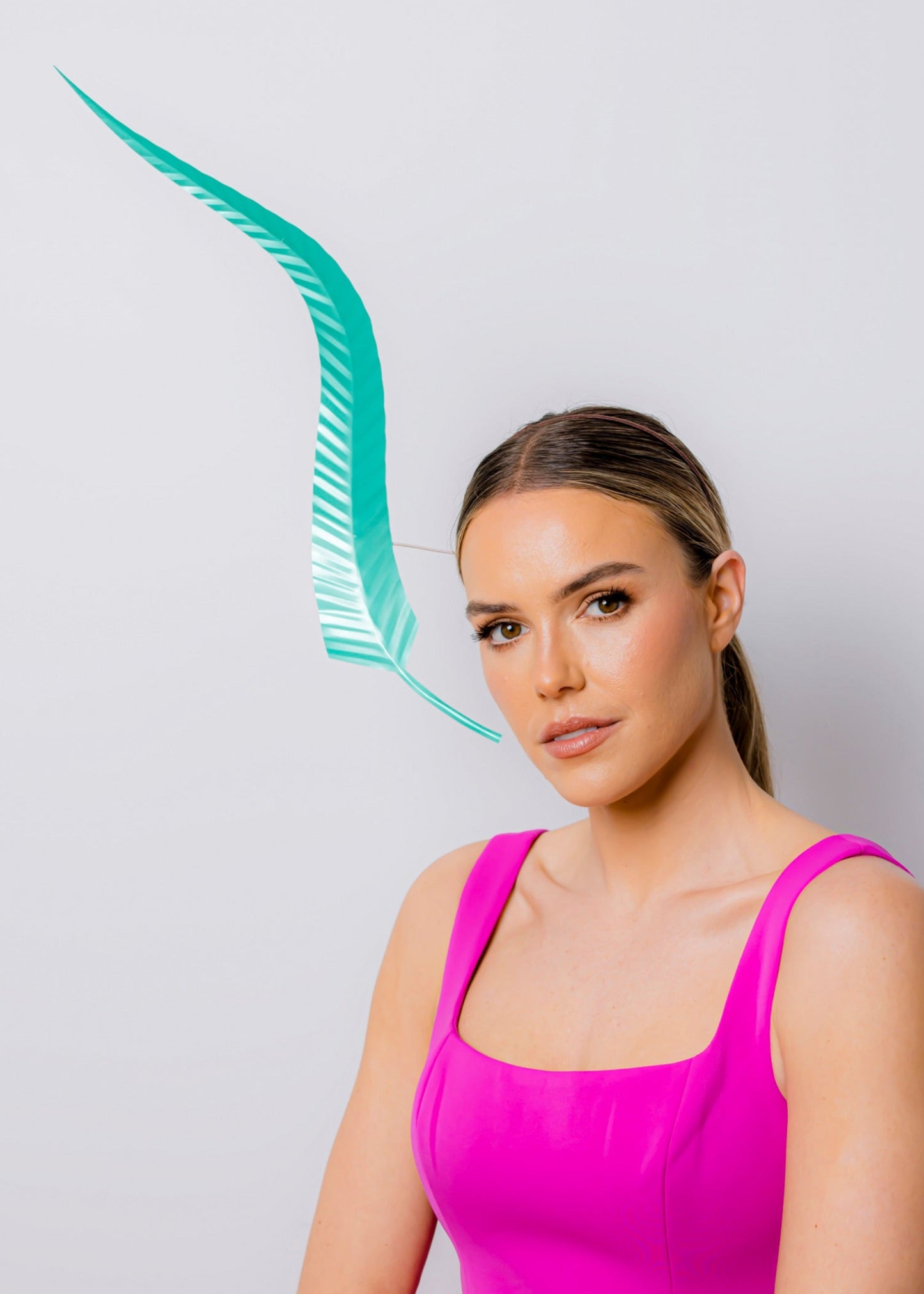 Woman in a pink dress standing against a light gray background with a green feather headpiece.