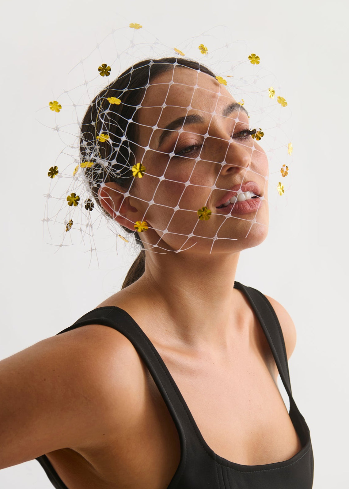 Woman wearing a black tank top with a white face veil and gold flowers on a light gray background