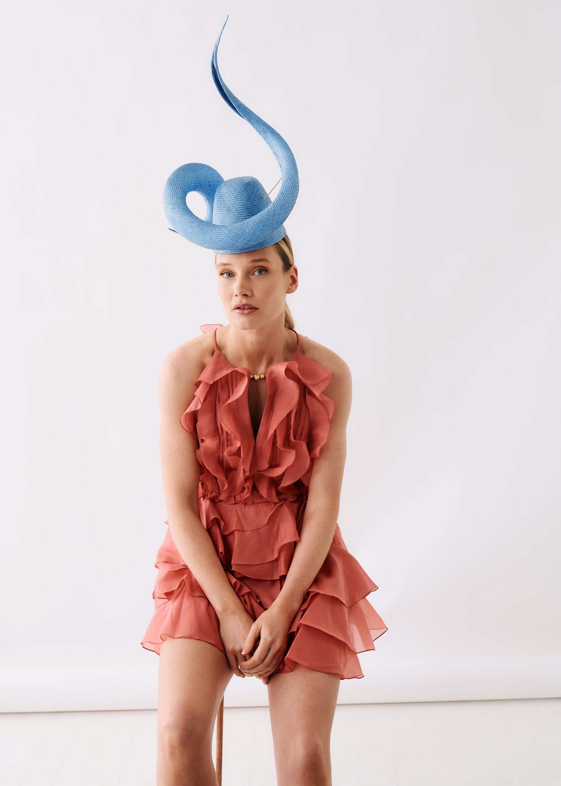 A model wearing a dusty rose ruffled halter mini dress and a large light blue fascinator hat, posed against a clean white studio background.