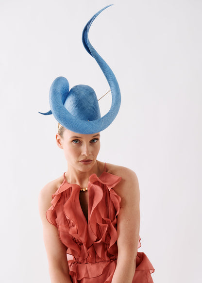 A model wearing a striking light blue fascinator hat with a long curved upward feather and a ruffled dusty rose halter dress, photographed against a clean white studio background.