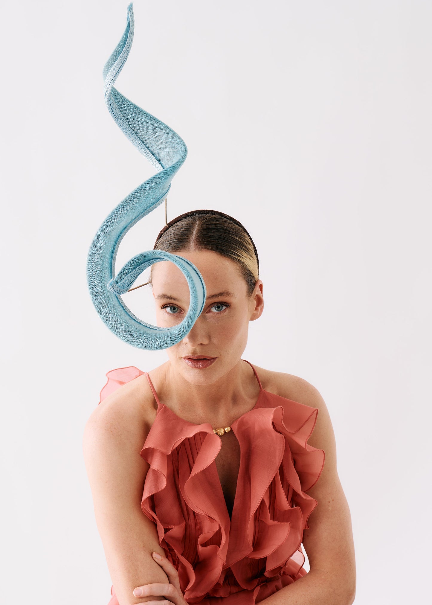 A model wearing a sculptural, flowing light blue fascinator headpiece and a ruffled coral pink dress against a plain light gray studio background.
