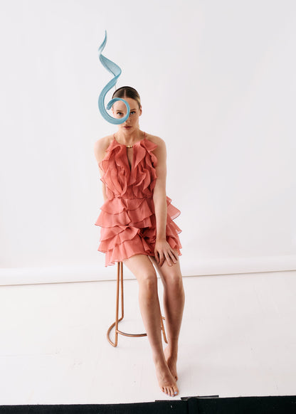 A model sits on a minimalist copper-toned stool in a studio setting, wearing a ruffled coral pink halter dress and a light blue sculptural headpiece.