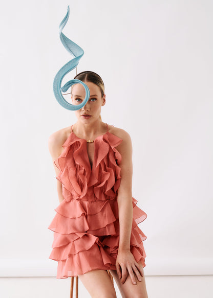 A model wearing a tiered ruffle halter dress in dusty rose pink, paired with a light blue sculptural fascinator headpiece, set against a plain white studio background.