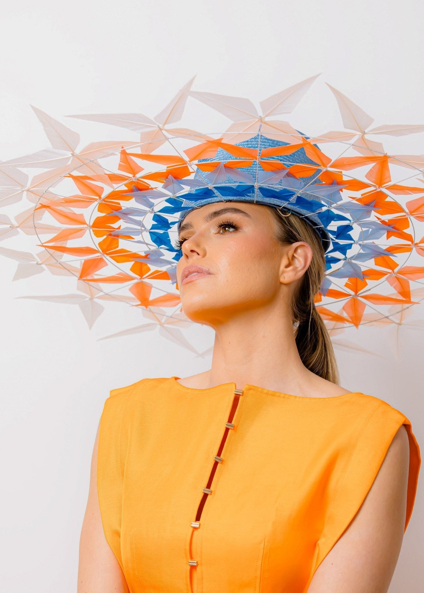 A woman wearing an orange dress and a PALOMA feather brimmed hat with blue and orange colors. The hat features a lattice effect design.
