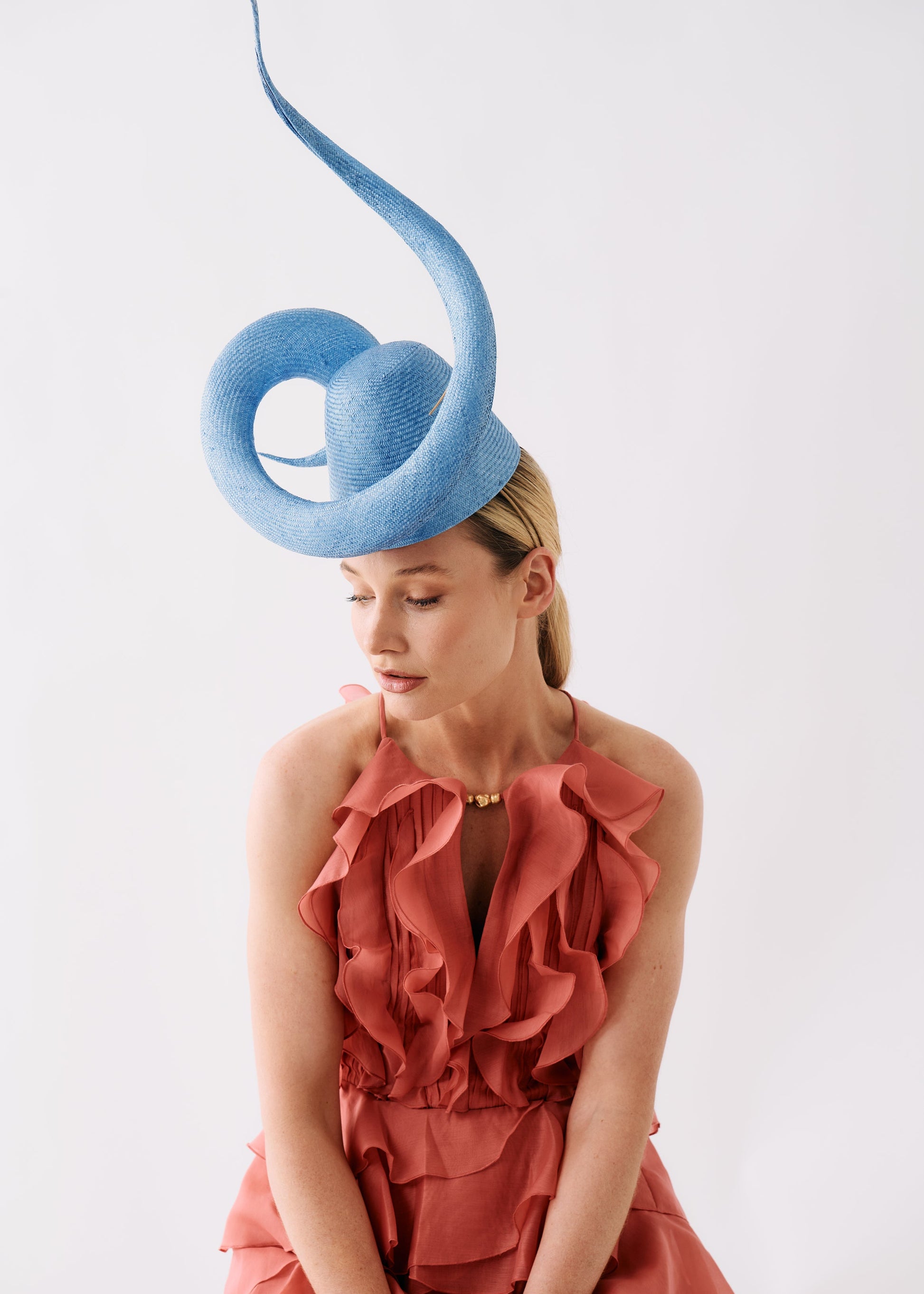 A woman wearing a dramatic light blue fascinator hat and a terracotta ruffled dress, seated against a plain white studio background.