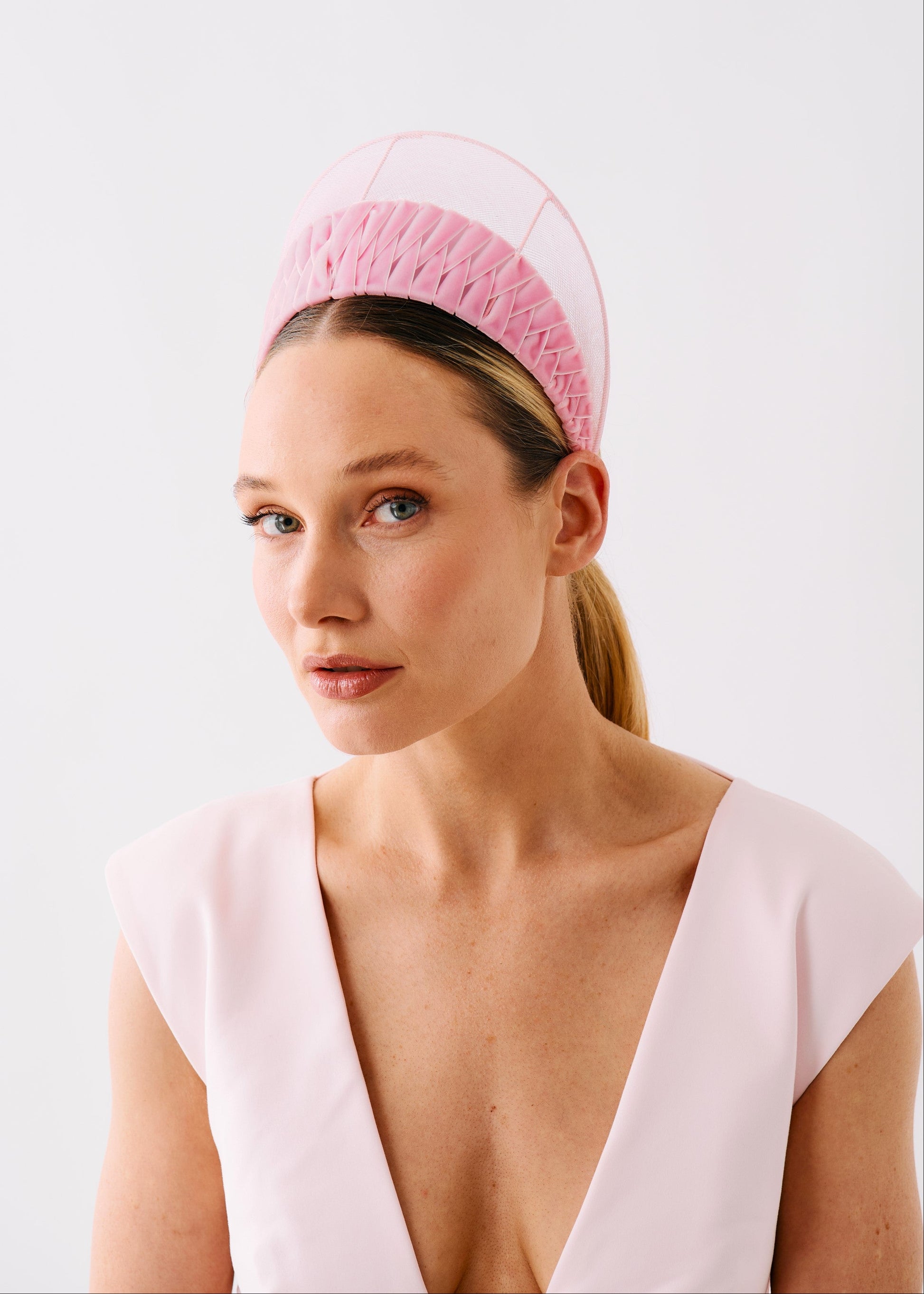 A woman wearing a structured light pink headpiece with a textured pink band and a pale pink upper section, paired with a pale pink sleeveless top against a plain light gray background.