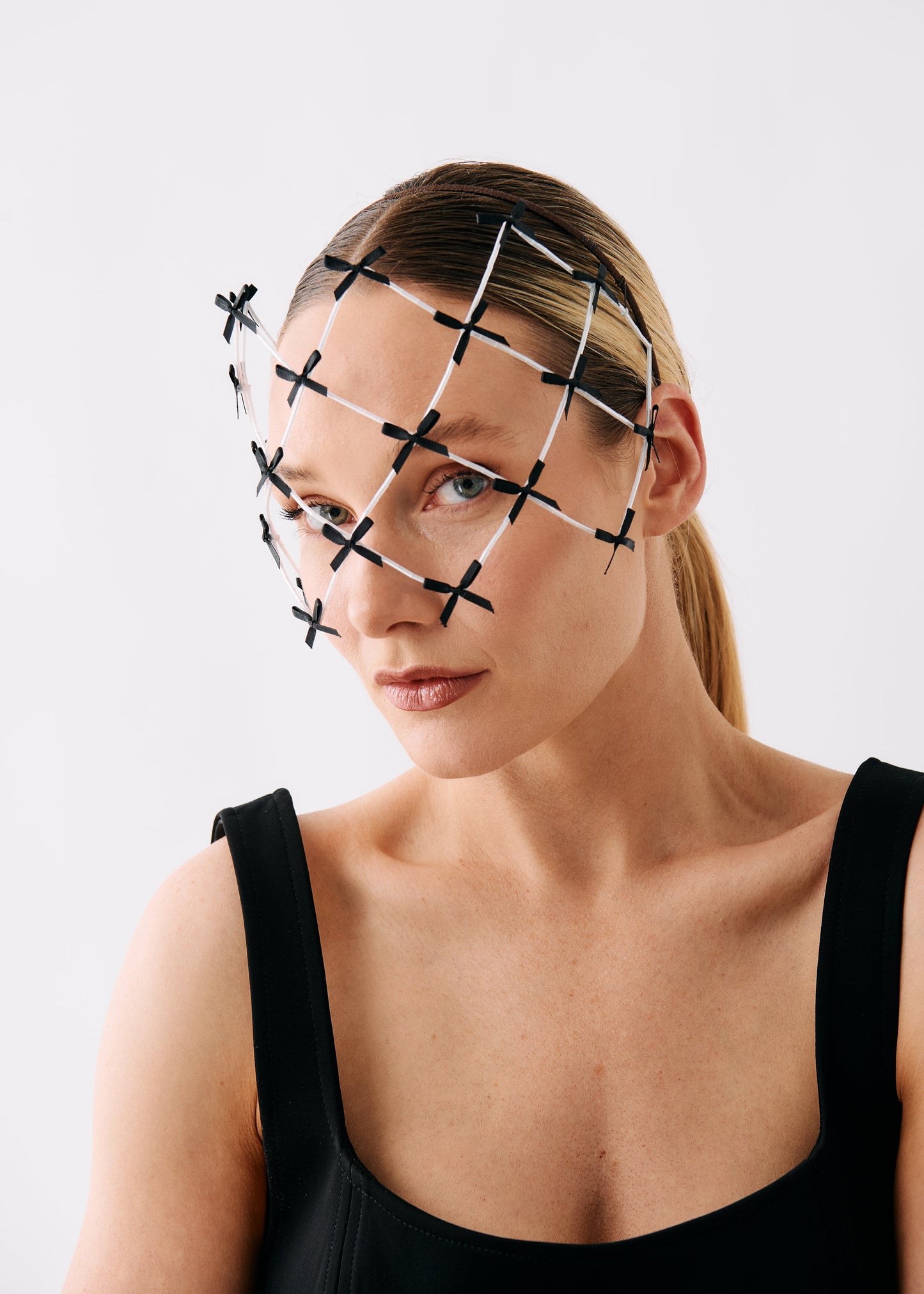 A woman wearing a black and white geometric wire fascinator headpiece over her eyes, paired with a black sleeveless top, photographed against a clean white studio background.