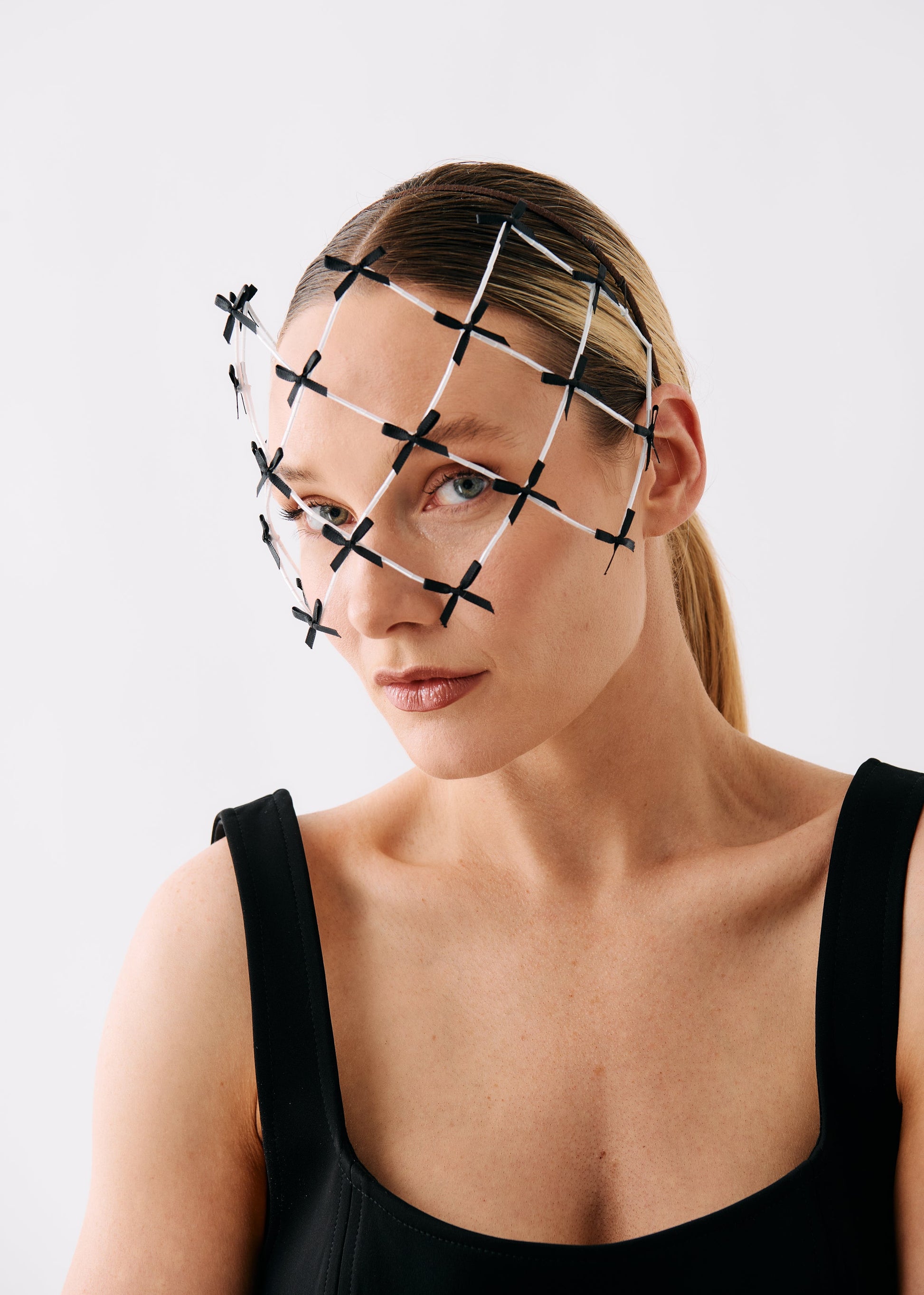 A woman wearing a black and white geometric wire fascinator headpiece over her eyes, paired with a black sleeveless top, photographed against a clean white studio background.