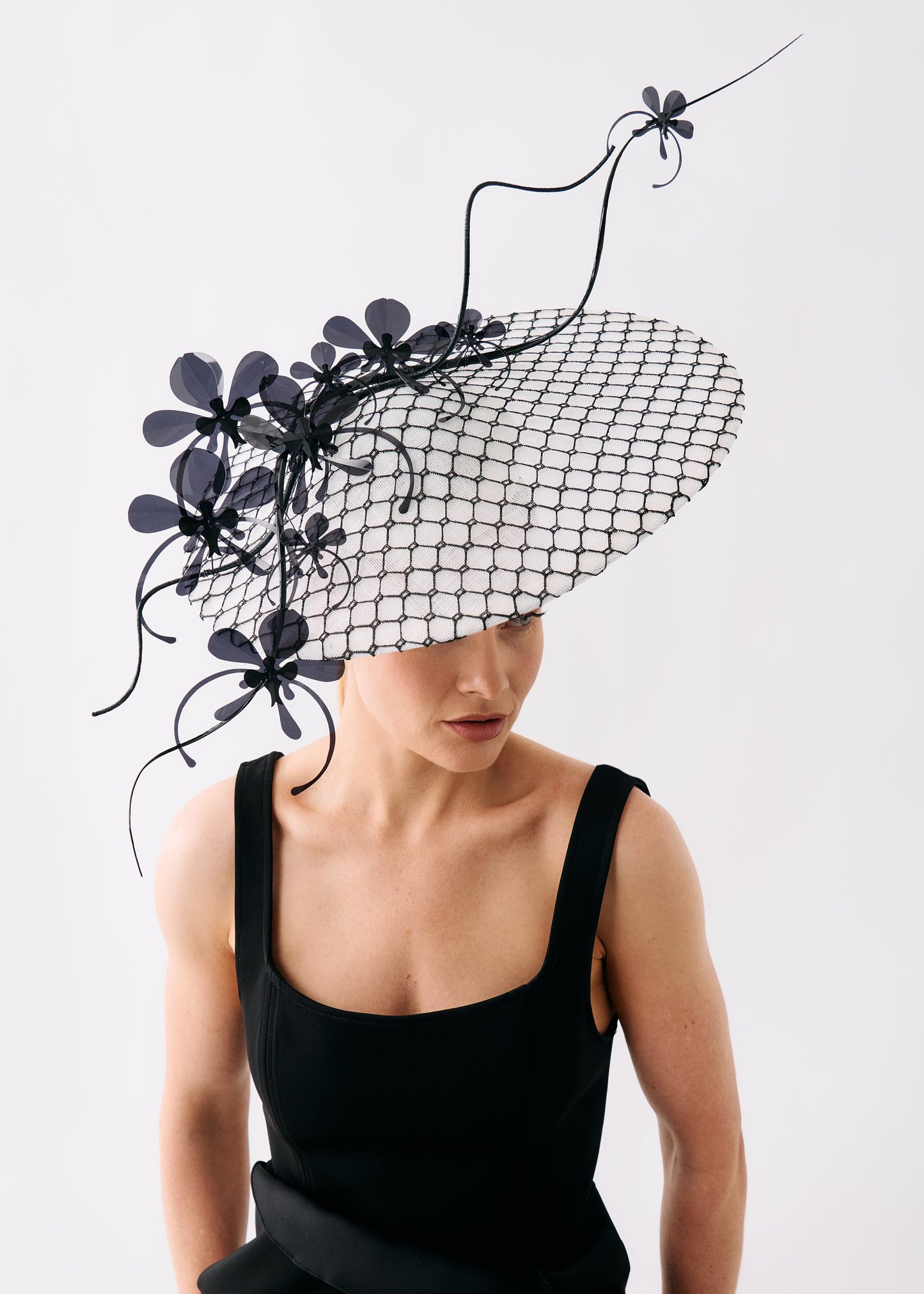 A model wearing a dramatic black fascinator hat with a black mesh brim, sculptural flower details, and curling black wire accents, paired with a black sleeveless dress against a plain light gray background.