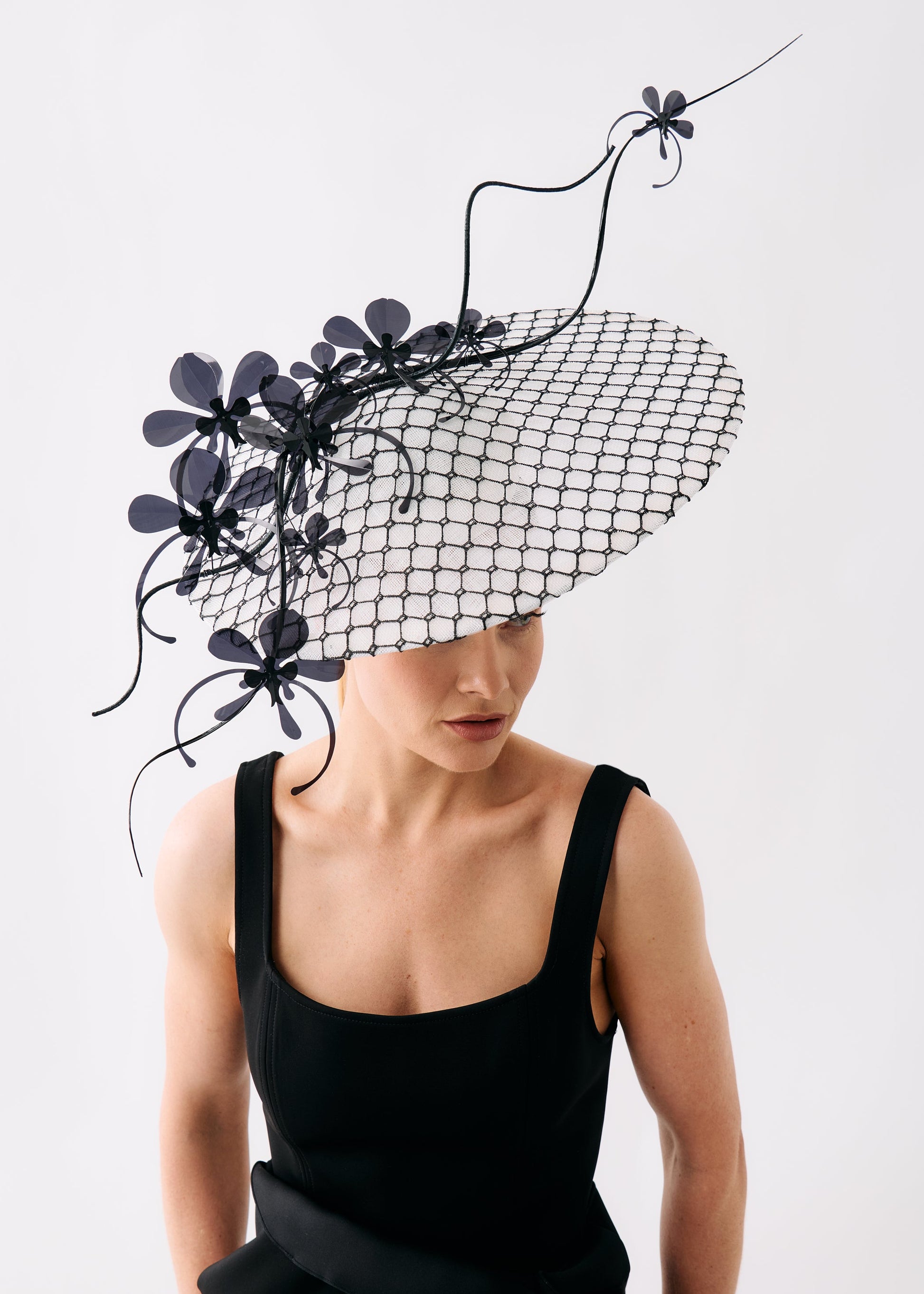 A model wearing a dramatic black fascinator hat with a black mesh brim, sculptural flower details, and curling black wire accents, paired with a black sleeveless dress against a plain light gray background.