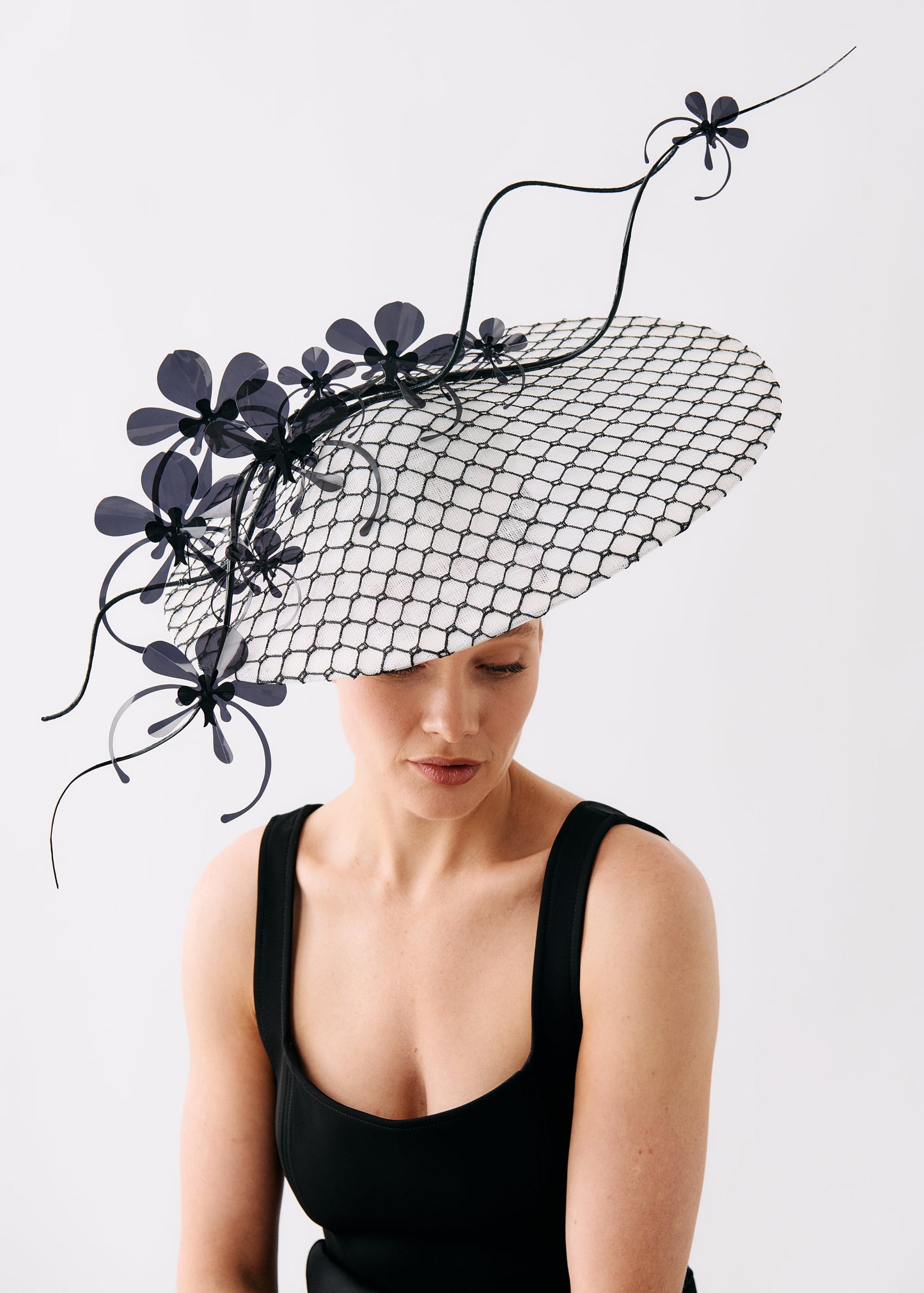 A woman wearing a large black fascinator hat with a black mesh brim, black floral accents, and curved black wire details, paired with a black sleeveless top, against a plain light gray background.