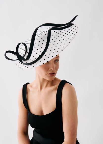 A woman wearing a large black-and-white polka dot fascinator hat with black ribbon trim, paired with a black sleeveless top, photographed against a light gray studio background.