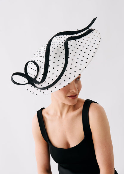 A woman wearing a wide-brimmed white hat with black polka dots and black ribbon trim, paired with a black sleeveless dress, photographed against a light gray studio background.