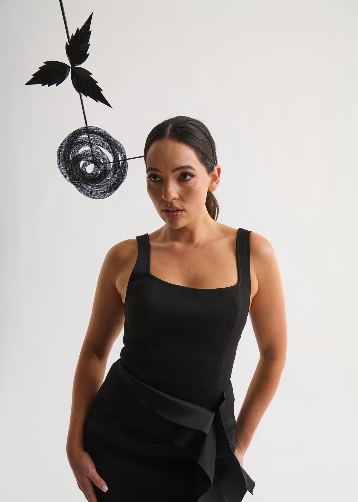 Woman wearing a black sleeveless dress with a decorative black flower headpiece against a white background