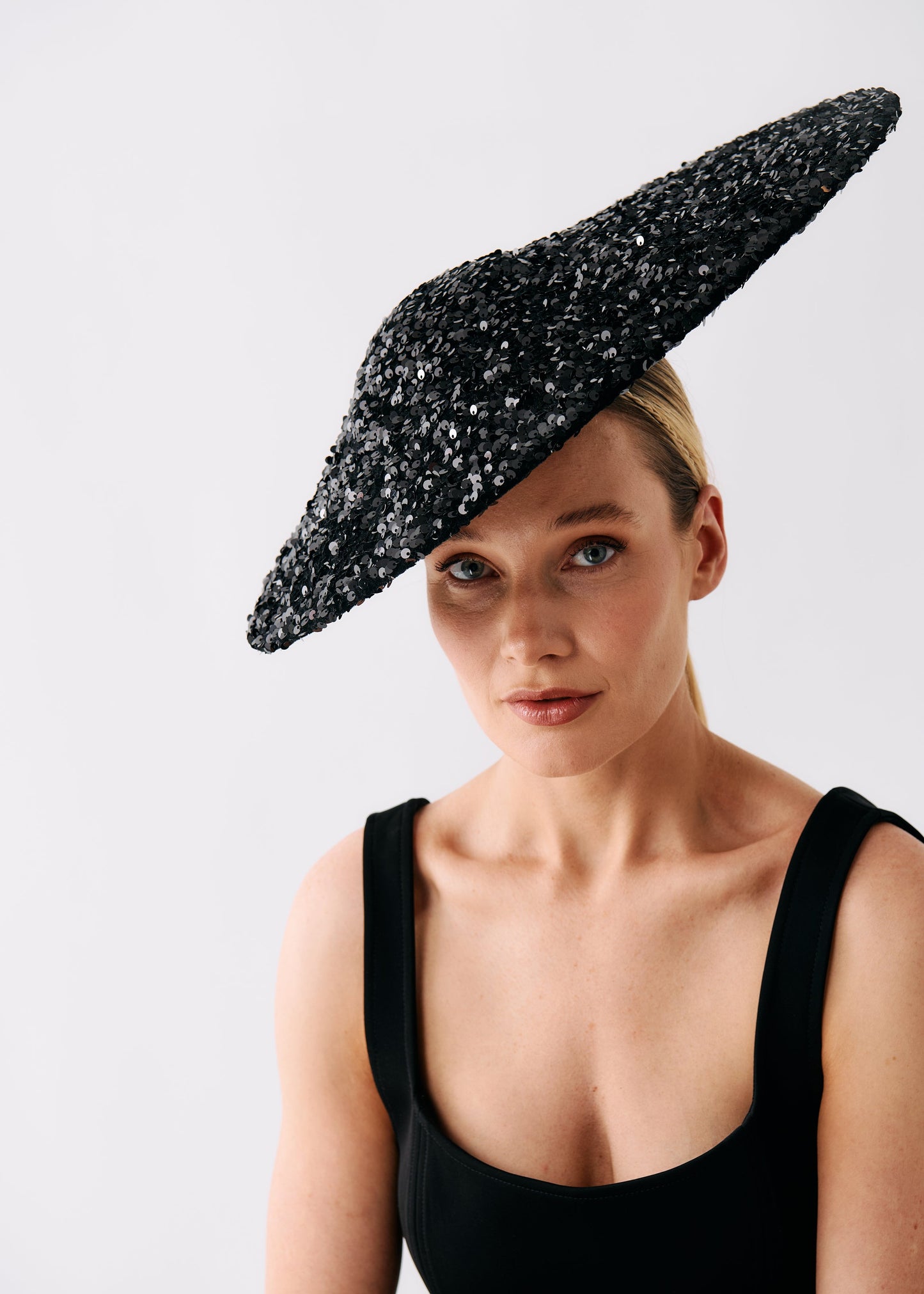 A woman wearing a dramatic black fascinator hat embellished with silver sequins, paired with a black sleeveless top, photographed against a light gray studio background.