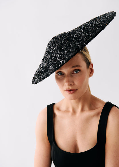A woman wearing a dramatic black fascinator hat embellished with silver sequins, paired with a black sleeveless top, photographed against a light gray studio background.
