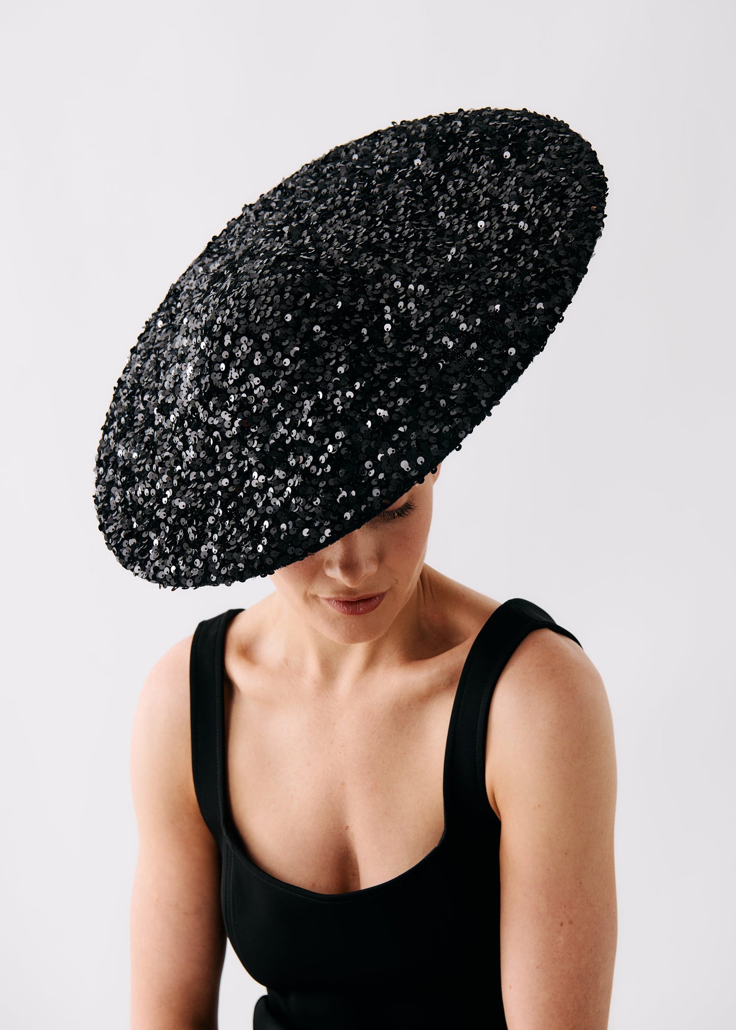 A woman wearing a large, wide-brimmed black fascinator hat covered in shimmering sequins, paired with a black sleeveless top, photographed against a clean light gray background.