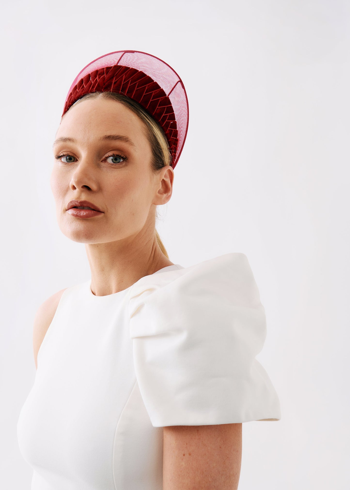 A woman wearing a structured white dress with dramatic puffed shoulders and a red mesh fascinator headpiece, photographed against a clean white studio background.