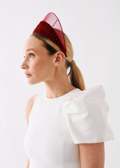 A woman in profile wearing a structured white dress with dramatic puffed sleeves and a red mesh fascinator headpiece, photographed against a clean white studio background.