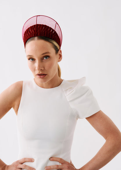 A woman wearing a structured white sleeveless dress with ruffled shoulders and a dark red mesh fascinator headpiece, posed against a light gray studio background.