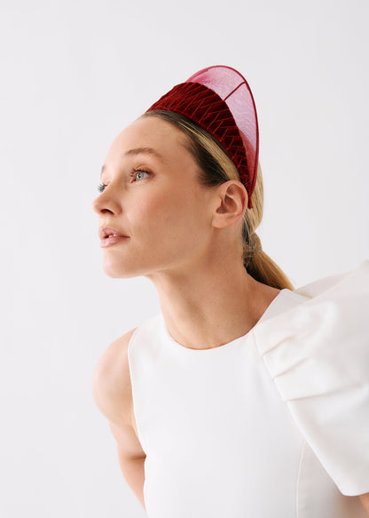 A woman in a white sleeveless dress wearing a structured burgundy velvet fascinator headpiece with a sheer pink mesh top, photographed against a clean white studio background.