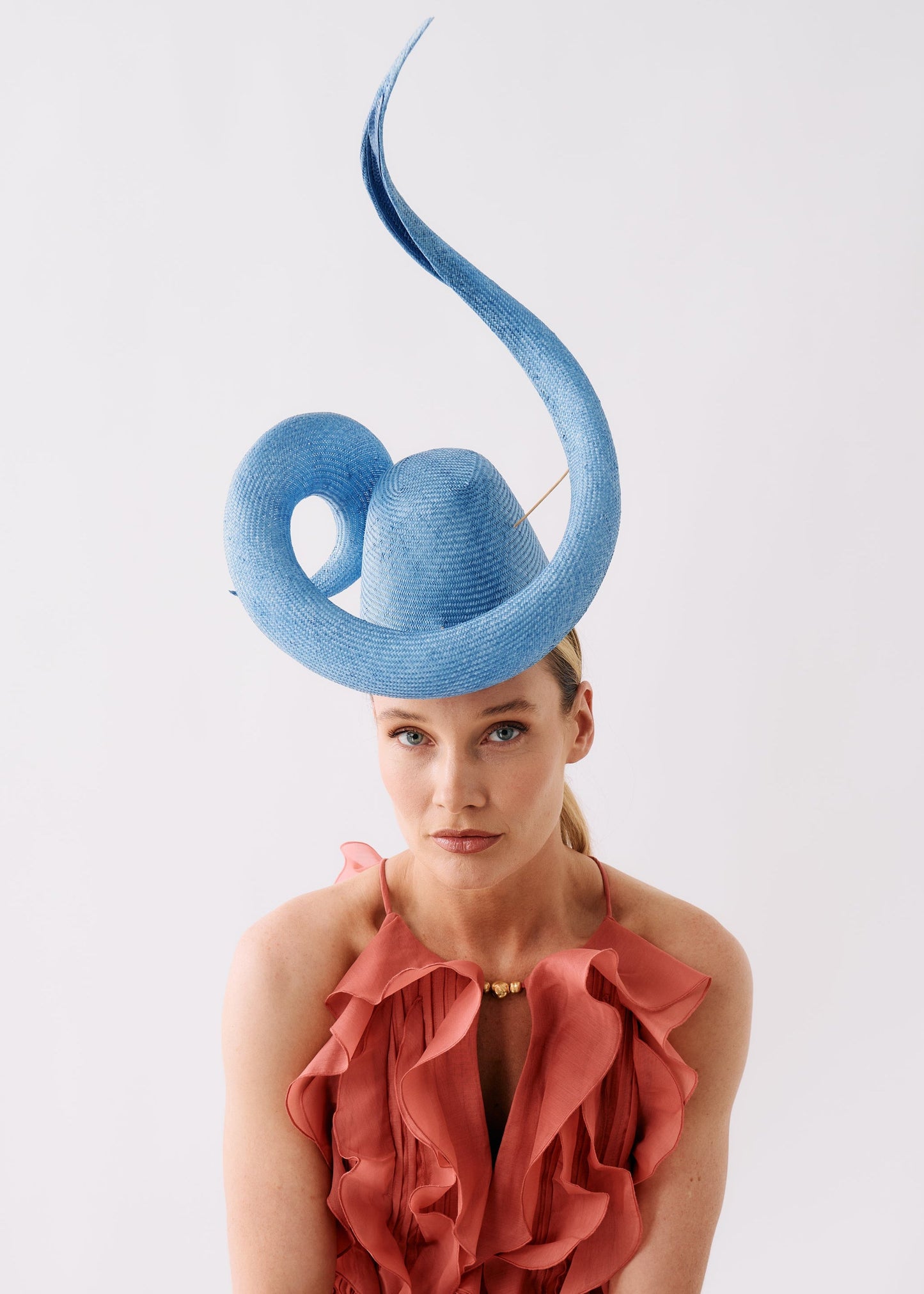 A model wearing a light blue sculptural fascinator hat and a ruffled dusty rose dress, photographed against a clean white studio background.