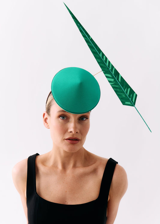 A model wearing a black sleeveless top and a distinctive green fascinator hat consisting of a round green dome with a long, leaf-shaped green feather attached, set against a plain light gray background.