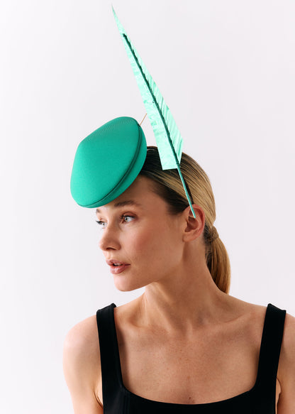 A woman wearing a bright green fascinator hat with a tall matching feather, photographed against a plain light background.