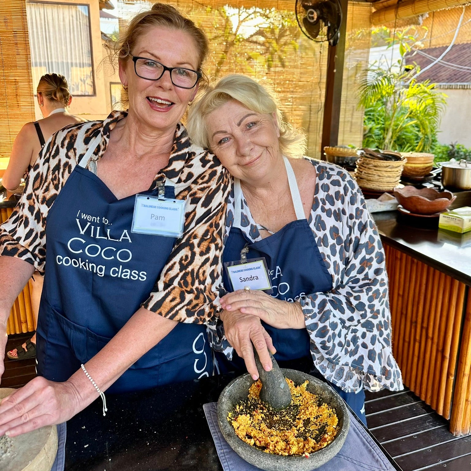 Two women in aprons at a cooking class, one holding a pestle and mortar with a bowl of ingredients.