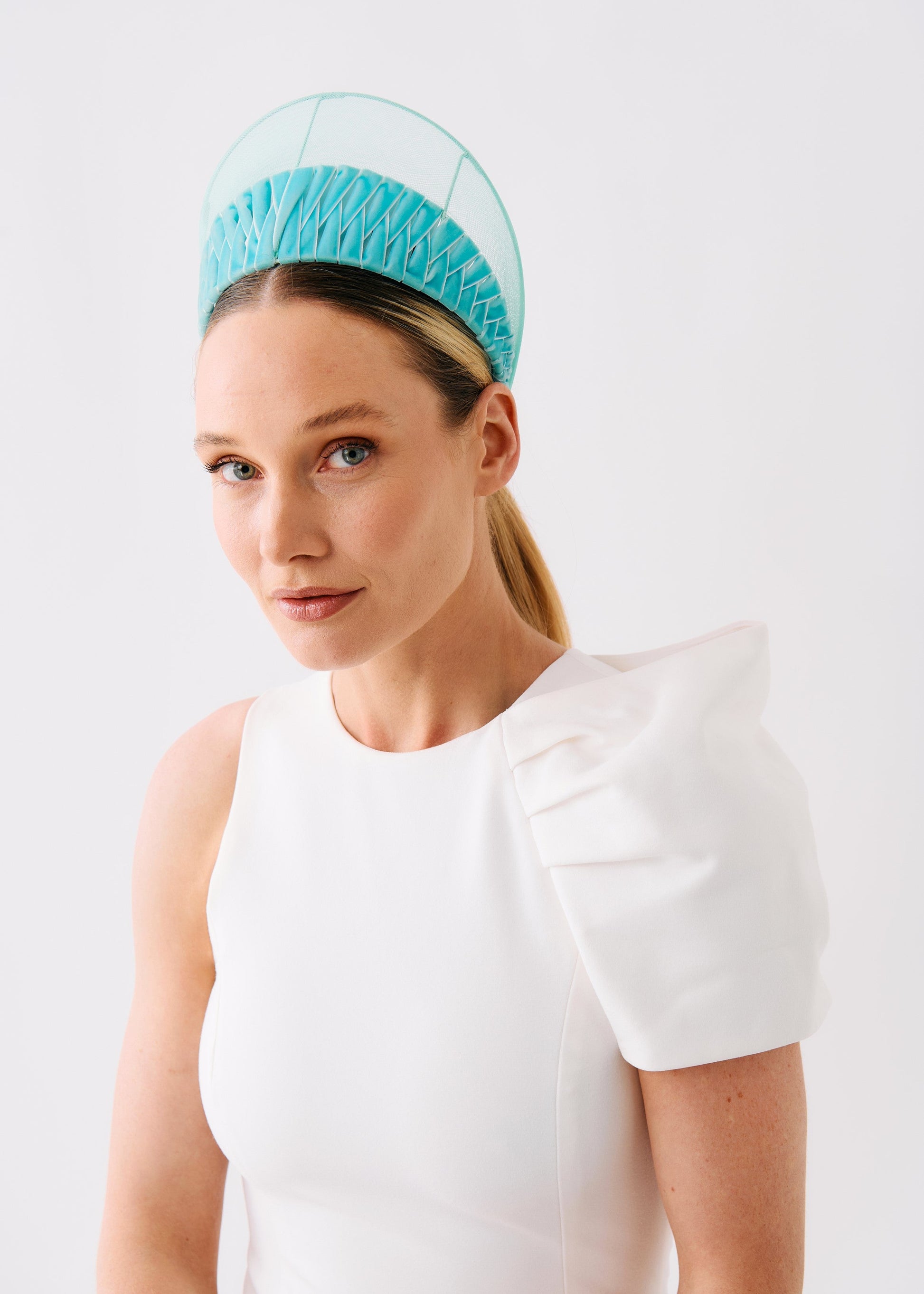 A woman wearing a white sleeveless dress with a dramatic puffed shoulder detail and a sheer light blue woven headband, photographed against a clean white studio background.