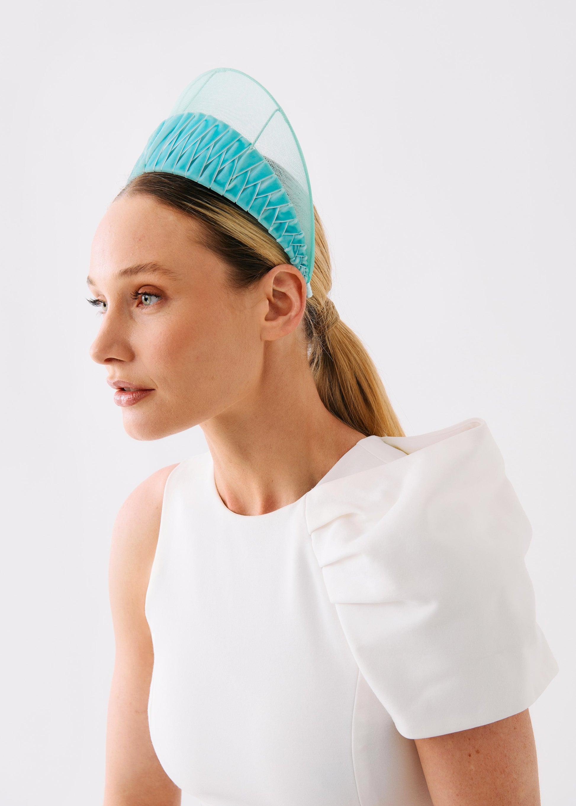 A woman in a white dress wearing a light blue pleated headband with a sheer mesh top, photographed against a plain white background.