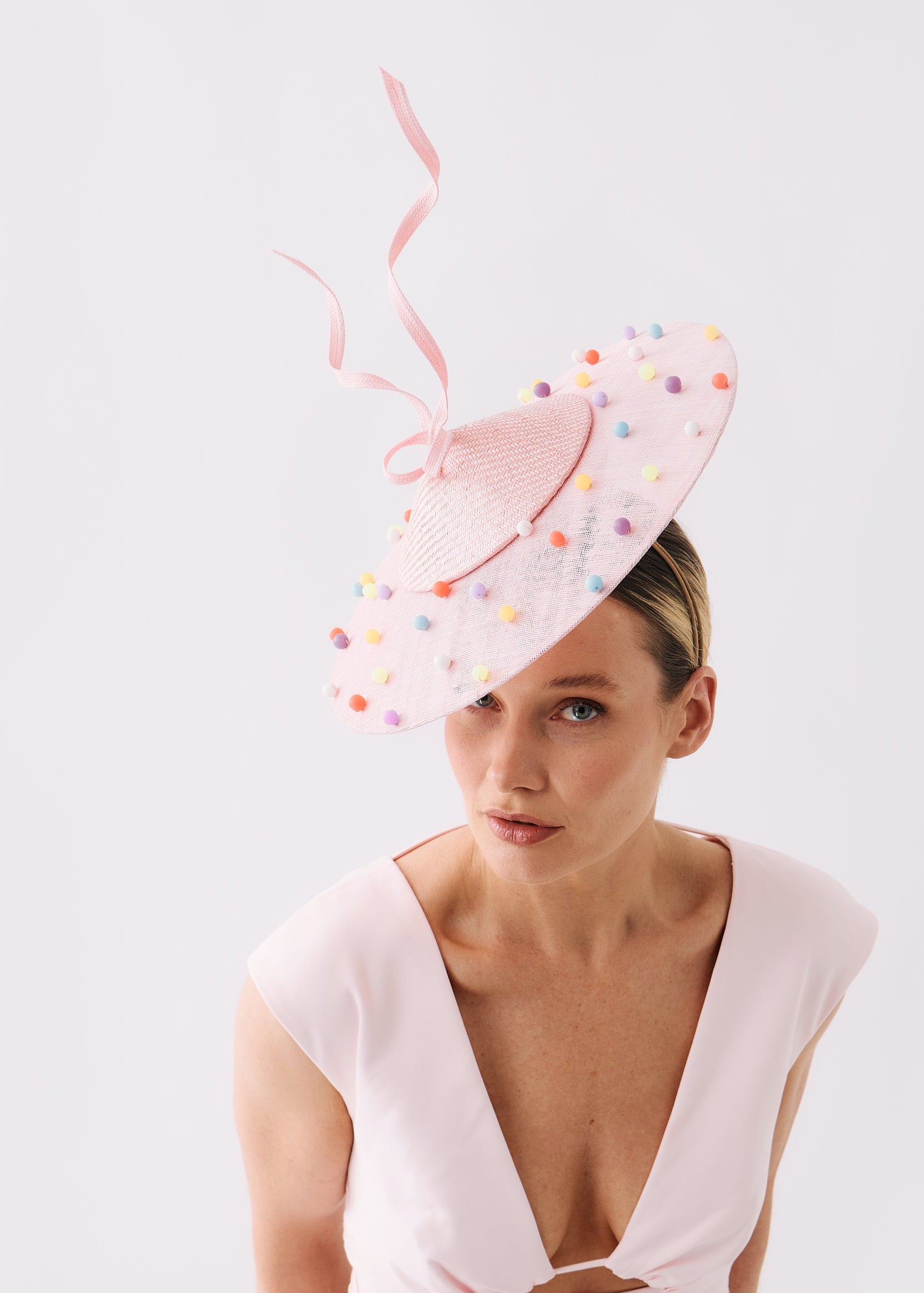 A woman wearing a pale pink fascinator hat decorated with colorful confetti dots and a long pink ribbon, paired with a matching pale pink dress against a plain white studio background.