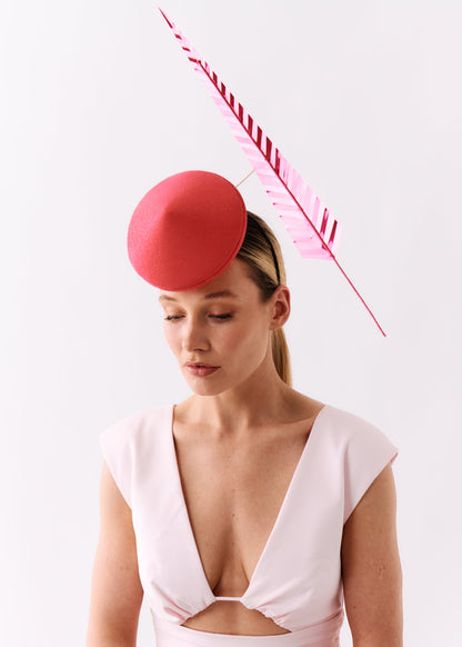 A woman wearing a pale pink dress and a bright pink fascinator hat with a matching pink feather, photographed against a clean white studio background.