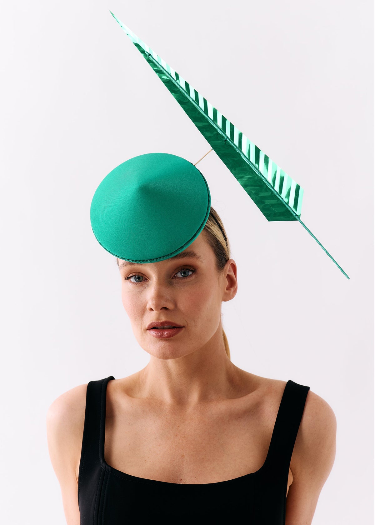 A woman wearing a black sleeveless top and a bright green fascinator hat with a matching green feather, photographed against a plain light gray studio background.