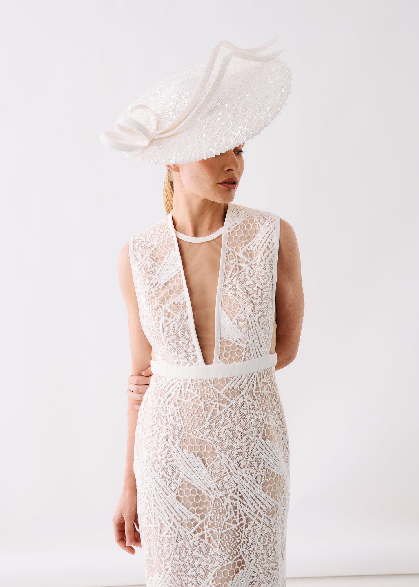 A woman wearing a sleeveless white lace dress with a deep V-neckline and a matching wide-brimmed fascinator hat, photographed against a clean white studio background.