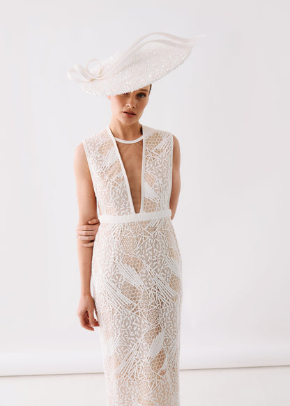 A model wearing a sleeveless, deep V-neck lace dress in cream/off-white with a matching waistband, paired with a large matching fascinator hat, against a plain white studio background.