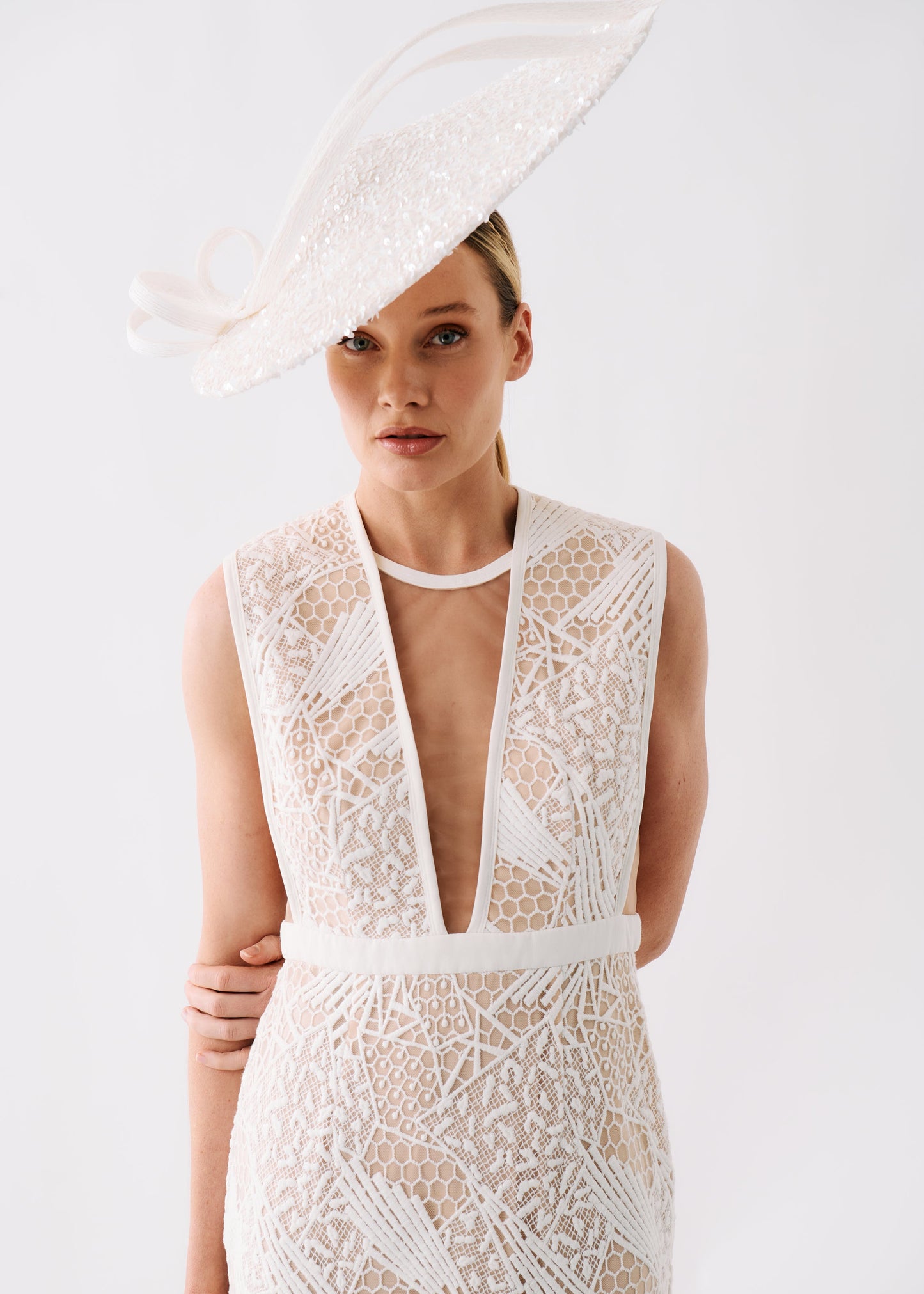 A woman wearing a sleeveless white lace dress with a deep V-neckline and a matching fascinator hat, photographed against a clean white studio background.