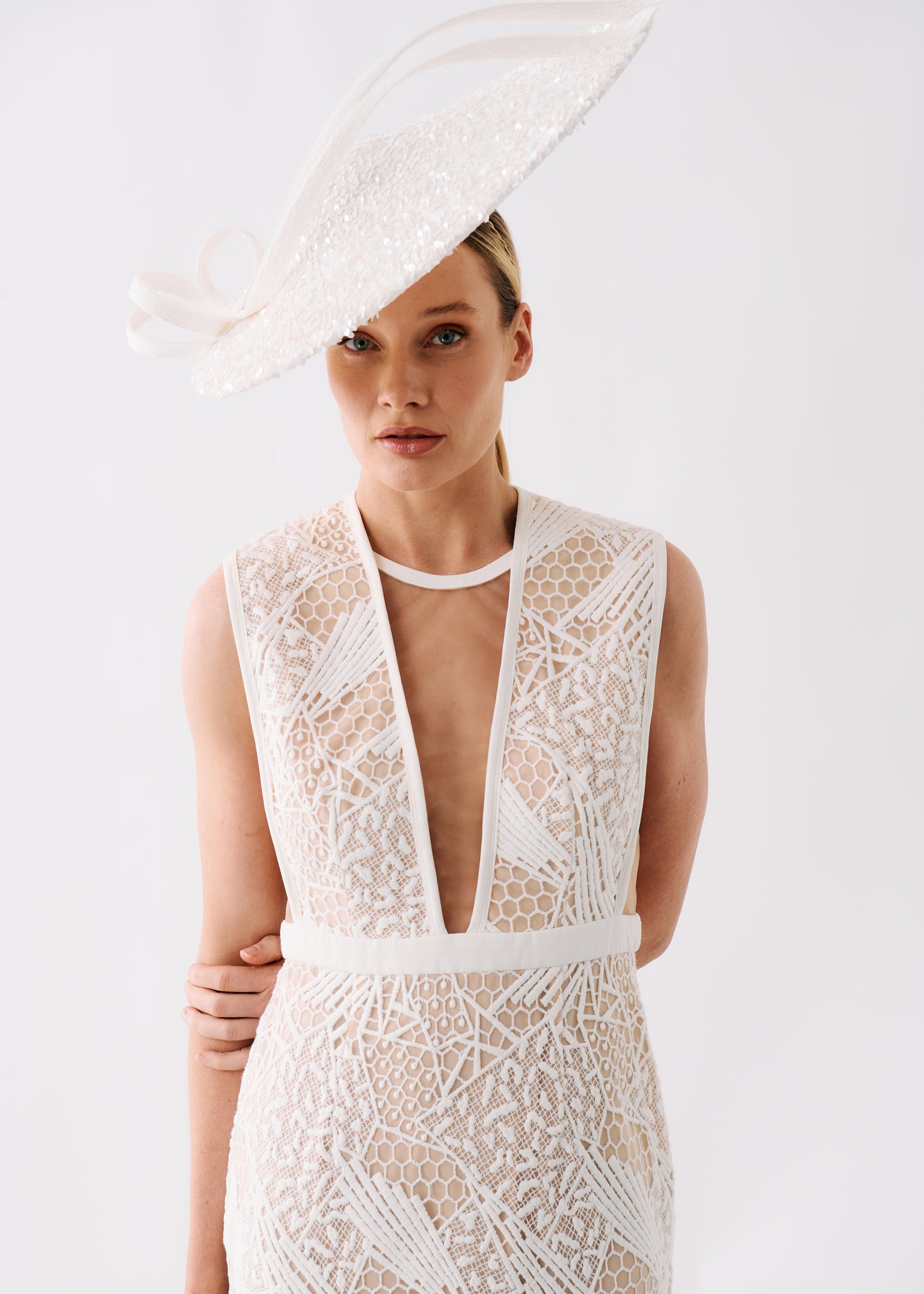 A woman wearing a sleeveless white lace dress with a deep V-neckline and a matching fascinator hat, photographed against a clean white studio background.