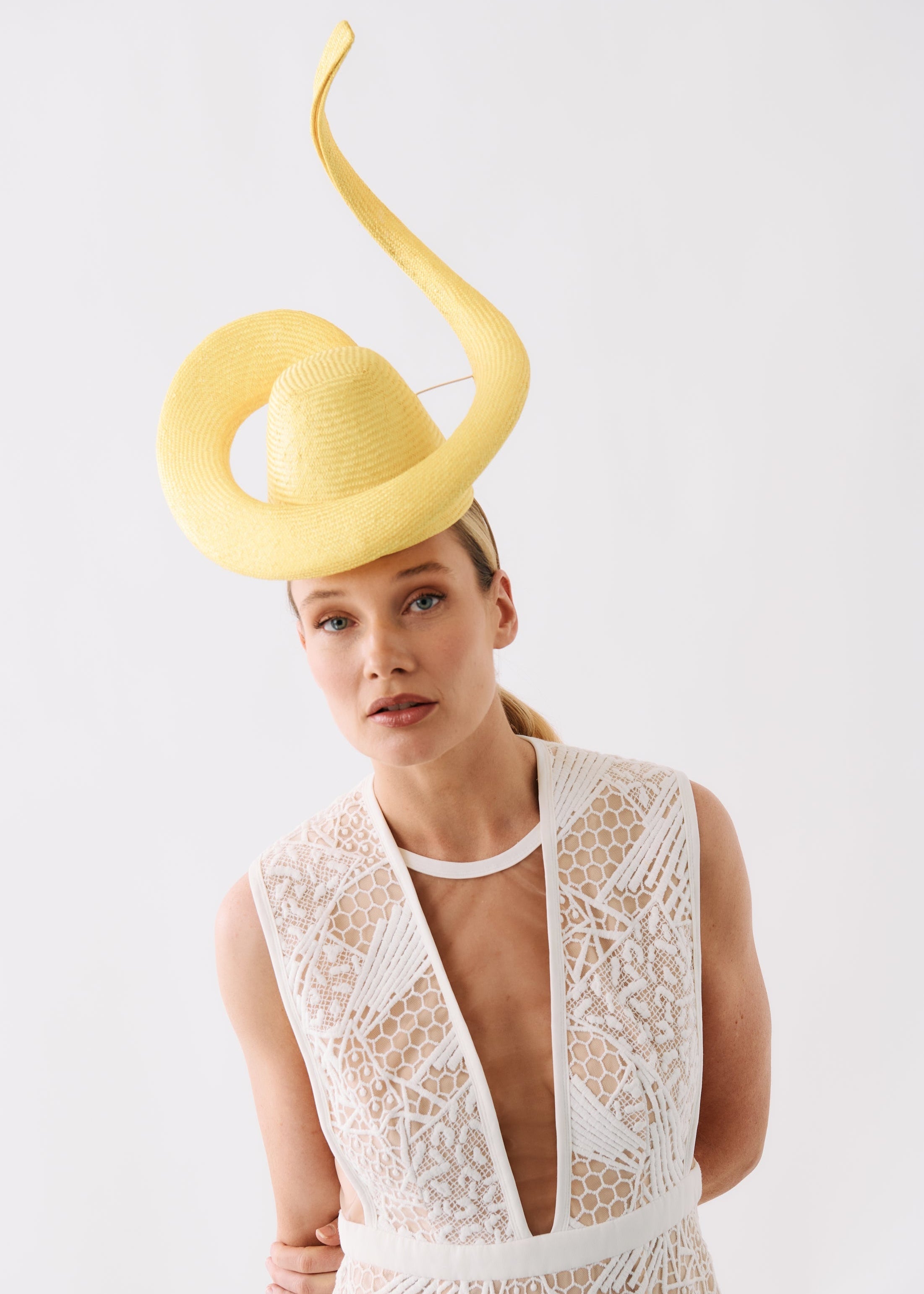A woman wearing a pale yellow fascinator hat with a long curved tail and a white lace dress, photographed against a plain light gray studio background.