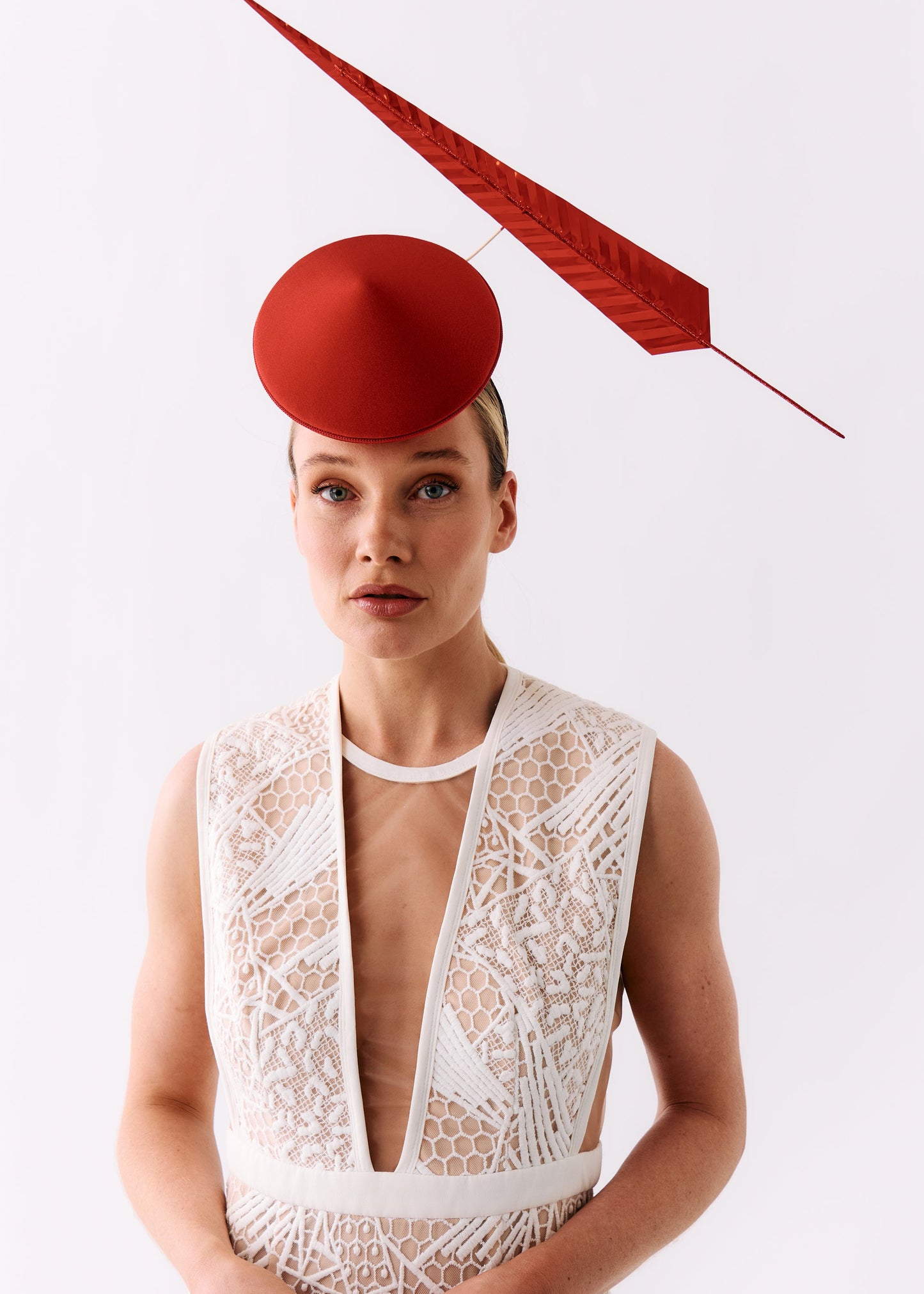 A model wearing a white lace dress with a deep plunging neckline and a matching white lace belt, styled with a red fascinator hat and a red feather headpiece against a white studio background.