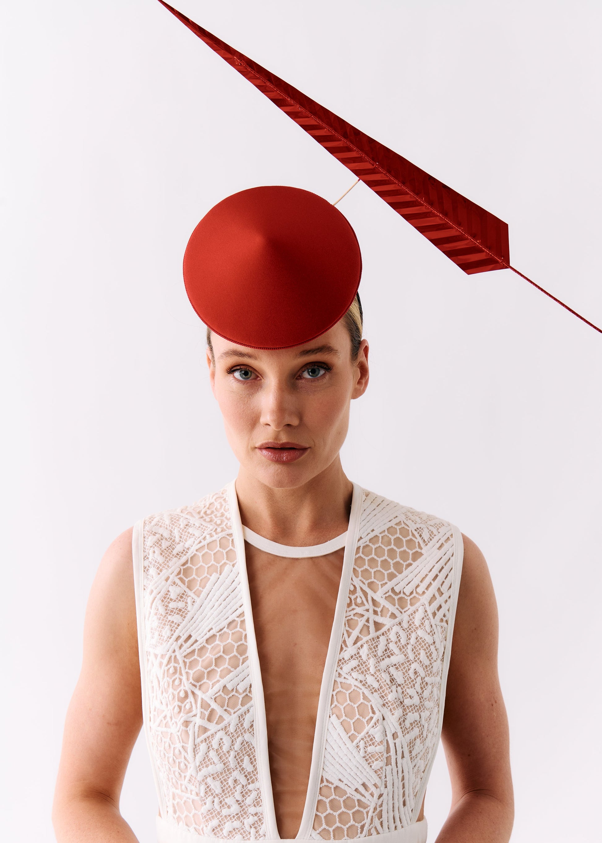 A model wearing a structured red fascinator hat and a white lace sleeveless top against a clean white studio background.