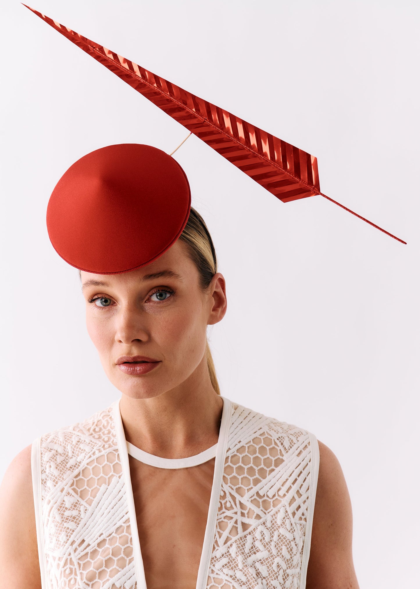 A model wearing a structured red fascinator hat and a white lace vest against a plain light gray background.