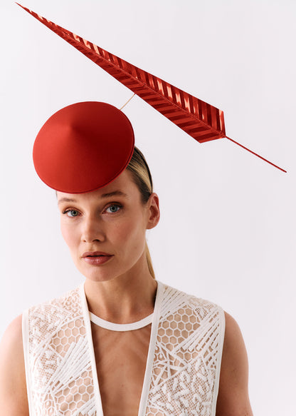 A model wearing a structured red fascinator hat and a white lace vest against a plain light gray background.
