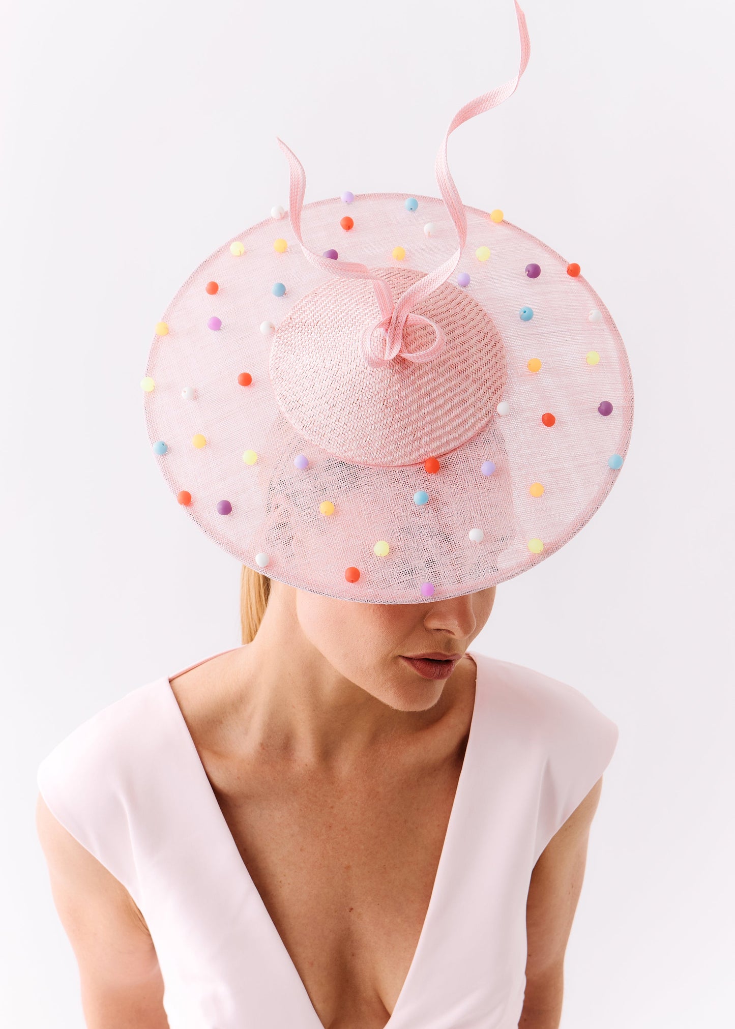 A woman wearing a pale pink wide-brimmed fascinator hat decorated with multicolored pom-poms and a matching pink ribbon, paired with a light pink sleeveless dress against a plain white background.
