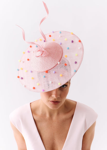 A woman wearing a pale pink wide-brimmed fascinator hat decorated with colorful beads and a matching pink ribbon, paired with a light pink dress against a plain white background.
