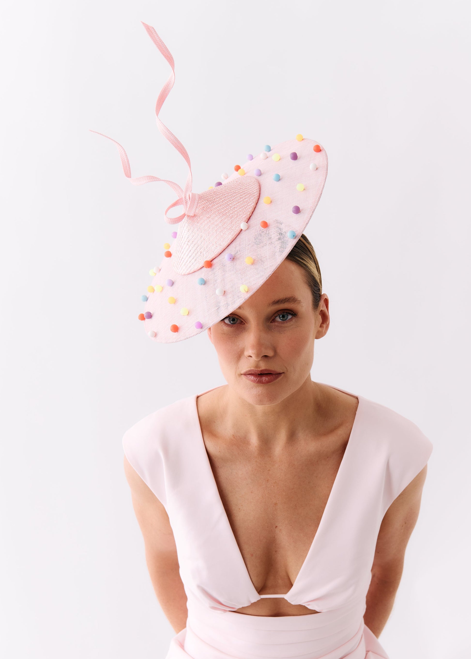 A woman wearing a pale pink dress and a large light pink fascinator hat decorated with small multicolored beads and ribbons, photographed against a clean white studio background.